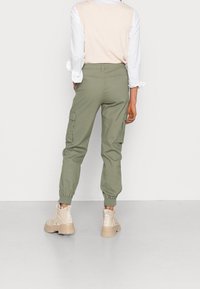 Olijfgroene cargobroek met elastische boorden, voorzien van zijzakken en een ontspannen pasvorm, gecombineerd met lichtbeige enkelboots en een gelaagde top.