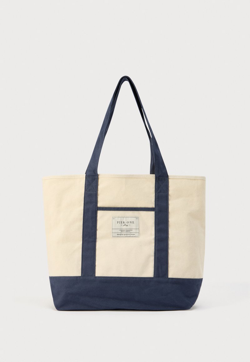 Sac fourre-tout en toile avec corps beige, fond et anses bleu marine. Présente une étiquette logo à l'avant avec des accents de couture. Forme rectangulaire.