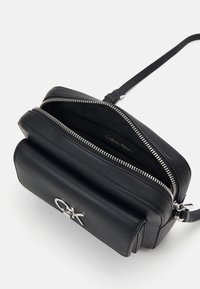 Schwarze Leder-Crossbody-Tasche mit einem Reißverschlussfach, silbernen Beschlägen und einem geprägten Calvin Klein-Logo auf der Vorderseite.