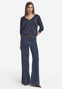 Navy Satinbluse mit V-Ausschnitt und Dreiviertelärmeln, kombiniert mit hochtaillierten, ausgestellten dunklen Jeans und einem braunen Gürtel.