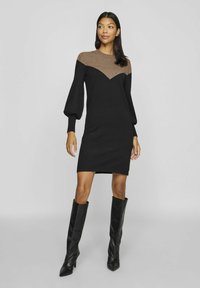 Robe en tricot noire avec un motif en chevron marron au niveau des épaules, manches bouffantes longues et ourlet droit. Assortie à des bottes à talons hauts noires.
