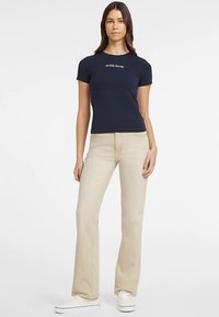 Tengerészkék pamut póló "GUESS JEANS" logóval, bézs színű bővülő szárú farmerrel és fehér tornacipővel párosítva. A modell egyenes testtartásban áll, semleges háttér előtt.