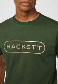Camiseta de algodón verde con cuello redondo y un logo dorado "HACKETT" impreso en un diseño rectangular en el pecho.