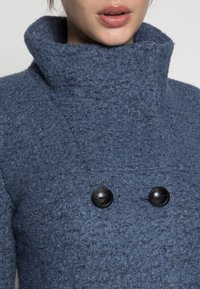 Modrý vlněný kabát s vysokým límcem, texturovaným povrchem, dvěma černými knoflíky a moderním designem.