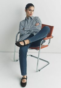 Des chaussures Mary Jane noires avec une sangle et une boucle, associées à un jean en denim bleu et à une chemise rayée noir et blanc, assises sur une chaise en chrome.