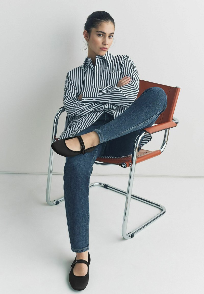 Pantofi Mary Jane negri cu curea și cataramă, asortați cu blugi albaștri și o cămașă în dungi alb-negru, așezată pe un scaun din crom.