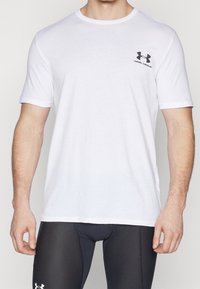 Camiseta blanca de manga corta hecha de algodón. Presenta un pequeño logotipo negro de "Under Armour" en el lado izquierdo del pecho. Diseño clásico de cuello redondo.