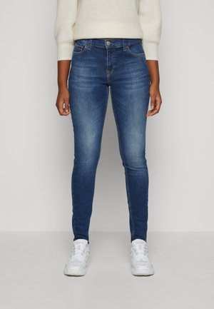 Jean skinny en denim bleu foncé avec une finition délavée. Comprend cinq poches, une fermeture à bouton et des coutures ton sur ton. Porté avec des baskets blanches.