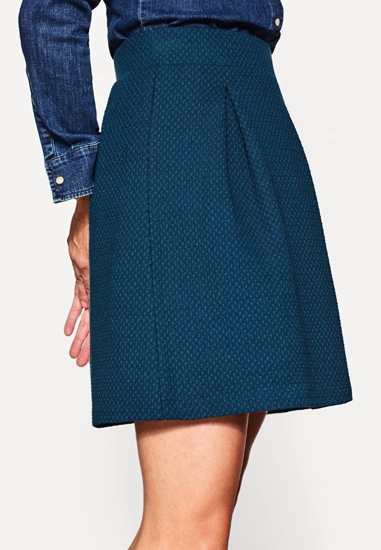 Femme portant une jupe bleue texturée longueur genou associée à une chemise en jean à manches longues, vue de la taille vers le bas, de profil.