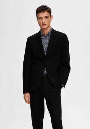 Traje negro a medida con solapas de muesca y bolsillos, usado sobre una camisa azul con estampado floral. Tejido suave con una silueta ajustada y afilada.