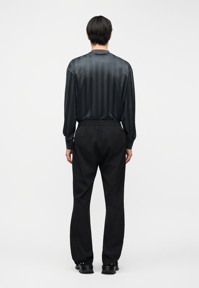 Άτομο με κοντά σκουρόχρωμα μαλλιά, φορώντας μαύρο μακρυμάνικο ριγέ πουκάμισο και φαρδιά μαύρη παντελόνια, να στέκεται γυρισμένο προς τα πίσω σε απλό φόντο.