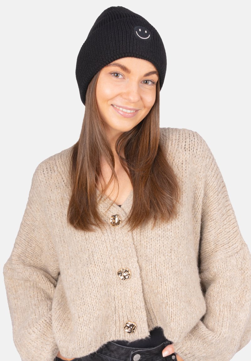 Bonnet en tricot noir avec un patch visage souriant blanc, porté avec un cardigan beige épais à gros boutons et une texture douce.