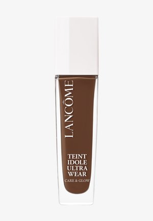 Flacon de fond de teint Lancôme, en verre rectangulaire avec un capuchon blanc, rempli d'un liquide brun riche. L'étiquette indique "Teint Idole Ultra Wear Care & Glow."