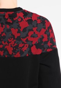 Felpa nera con una sezione superiore in tessuto camuffato rosso e nero. Collo a costine nero, materiali in contrasto e dettagli di design.
