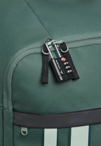 Zaino in tessuto verde con cerniera, dotato di una combinazione nera e dettagli in tessuto nero. Include strisce bianche verticali lungo il lato.