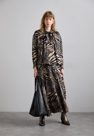 Pitkä, beige ja musta zebra-kuvioinen mekko, jossa on nauhakolla, yhdistettynä mustaan laukkuun ja metallinhohtoisiin nilkkureihin, seisten harmaalla lattialla.