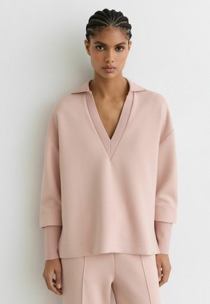 Frau mit geflochtenem Haar, die ein hellrosa V-Ausschnitt-Longsleeve und passende Hose trägt, steht vor einer schlichten weißen Wand.