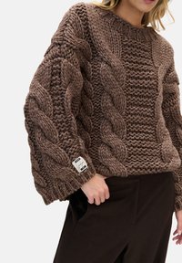 Brązowy sweter z grubym, plecionym wzorem, ściągaczami na mankietach i zaokrąglonym dekoltem. Metka z informacjami o marce na lewym rękawie.