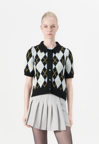 Černý kardigan s krátkým rukávem a argyle vzorem v světle modré a žluté, s límečkem a černými knoflíky. Stylizováno s plisovanou šedou sukní.