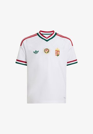 Dreng står med den ene hånd i lommen iført hvid sportsjersey med røde og grønne striber, vide blå jeans og sorte sko.