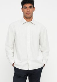 Uomo che indossa una camicia bianca a maniche lunghe con sottili righe verticali e pantaloni scuri, in piedi con le mani in tasca contro uno sfondo semplice.
