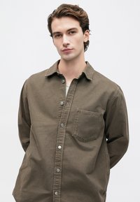 Joven con cabello castaño estilizado lleva una camisa de botones marrón sobre una camiseta blanca, de pie contra un fondo gris claro liso.