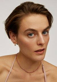 Aros de hojalata dorada y un collar de cuentas rosas con un colgante de la letra 'K'. El sujeto tiene el pelo corto y un tono de piel claro. Fondo sencillo.