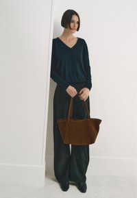 Bruine suède totebag met twee handvatten, trapeziumvormig, gecombineerd met een donker V-hals top en brede zwarte broek, geplaatst tegen een witte achtergrond.