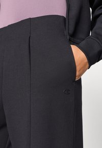 Μαύρο αθλητικό παντελόνι με υφή, διαθέτει πτυχώσεις μπροστά και πλαϊνές τσέπες. Ένα μικρό, σκοτεινό λογότυπο είναι ορατό στο κάτω μέρος του ποδιού.