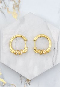 Orecchini a cerchio in oro con design intrecciato che presenta piccole pietre di strass trasparenti, su uno sfondo di marmo. Forma circolare con accenti testurizzati.
