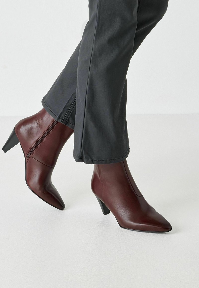 Bottes chevilles en cuir bordeaux avec un bout pointu et un talon de 7 cm, dotées d'une fermeture éclair latérale et d'une texture lisse, portées avec un pantalon gris.
