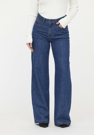 Wijde blauwe denim jeans met een hoge taille, zakken aan de voorkant en subtiele vervaging. Draag met een witte geribbelde top met lange mouwen.