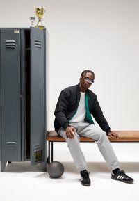 Μαύρο μπουφάν bomber με πράσινη επένδυση, γκρι φόρμα, μαύρα αθλητικά παπούτσια, σε ένα ξύλινο παγκάκι δίπλα σε γκρι ντουλάπι και τρόπαια. Μπάσκετ κοντά.