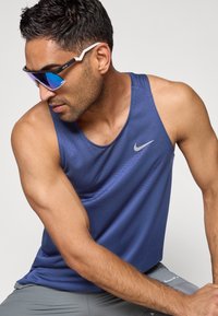 Maiou Nike bleumarin cu guler rotund și model subtil, asortat cu ochelari de soare eleganți de tip wrap, cu lentile albastre reflectorizante.