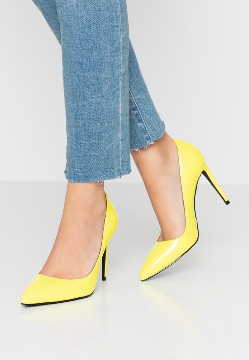 Jambes portant un jean bleu coupé et des chaussures à talons hauts en cuir verni jaune avec des bouts pointus sur une surface claire.