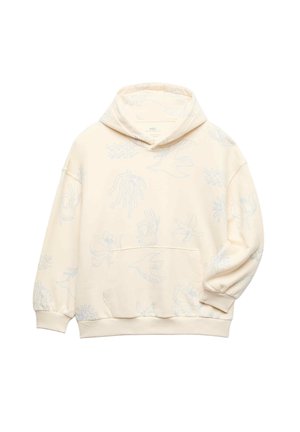 Ung person med kort hår iført en cremefarvet hættetrøje med diskrete mønstre, løse blå jeans og beige sneakers, stående med hænderne i lommerne.