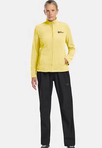 Veste polaire jaune avec col montant, fermeture éclair devant et poches latérales, associée à un pantalon noir. Design simple et texture douce.