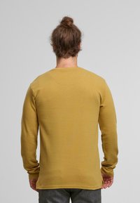 Hombre con cabello castaño recogido en un moño, vistiendo una camisa de manga larga color mostaza y pantalones grises, de espaldas frente a un fondo simple.