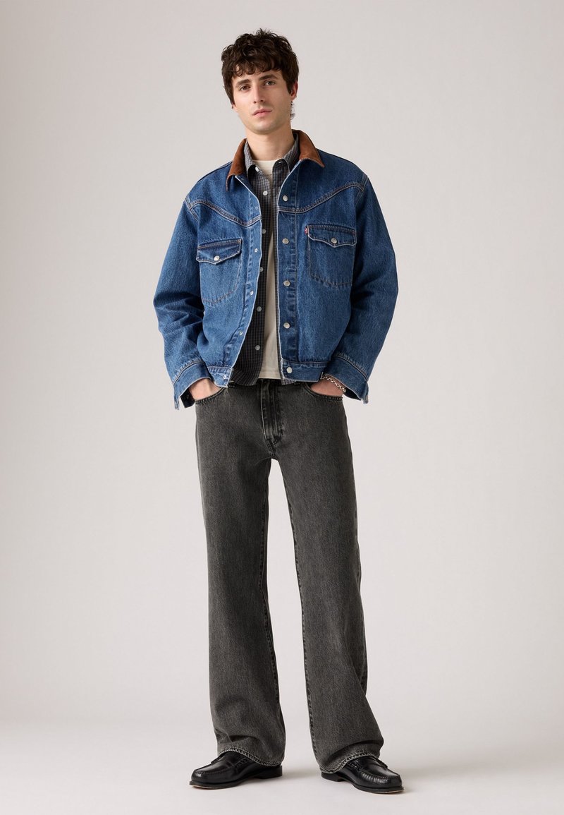 Jeune homme debout les mains dans les poches, portant une veste en denim bleu avec col marron, une chemise à carreaux, un jean gris et des chaussures noires.