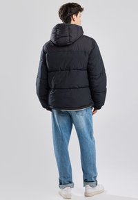 Black puffer jacket with a hood, featuring quilted sections and a smooth finish. Paired with light blue jeans and white sneakers.