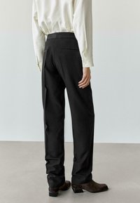 Schwarze Hose mit geradem Bein und Gesäßtaschen, kombiniert mit einem locker sitzenden, langärmligen weißen Hemd und braunen Knöchelstiefeln.