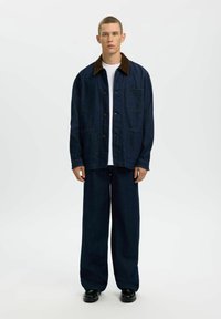 Homme debout portant une veste en denim bleu foncé avec un col marron, un jean large assorti, une chemise blanche et des chaussures noires sur un fond blanc.