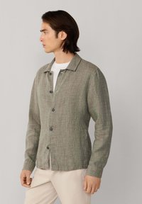 Muž v zapnutom šedej zelenkavej textúrovanej bunde cez bielu košeľu a svetlobéžové nohavice, otočený doľava na jednofarebnom pozadí.