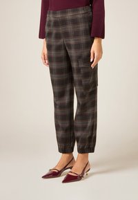 Geruite broek in donkere tinten met bruine en lichtgrijze strepen. Elastische boorden en een zijzak zorgen voor extra functionaliteit. Draagt bijpassende bordeauxrode hakken.