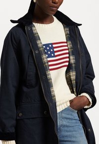 Abrigo azul marino con collar de pana, forro de cuadros; suéter crema con un gráfico de la bandera americana en rojo y azul; jeans de mezclilla claros.