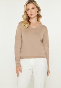 Beige V-hals trui met lange mouwen, geribde manchetten en zoom, gemaakt van zacht materiaal. Gecombineerd met witte broeken. Soepele textuur.
