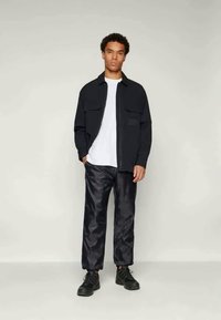 Musta takki, jossa on kaksi etutaskua, valkoisen T-paidan päällä. Yhdistetty kiiltäviin tummiin housuihin ja mustiin lenkkareihin.