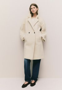 Double-breasted cream coat with a textured wool-like material, oversized shape, and black buttons; worn over loose blue jeans and dark shoes.