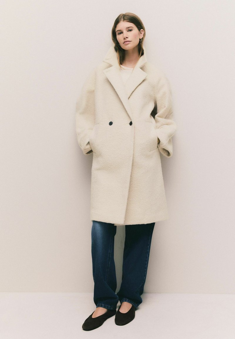 Double-breasted cream coat with a textured wool-like material, oversized shape, and black buttons; worn over loose blue jeans and dark shoes.
