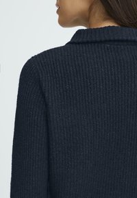 Tengerészkék kötött pulóver bordázott textúrával, hajtókás gallérral és szabott fazonnal, amely letisztult és modern dizájnt mutat be.